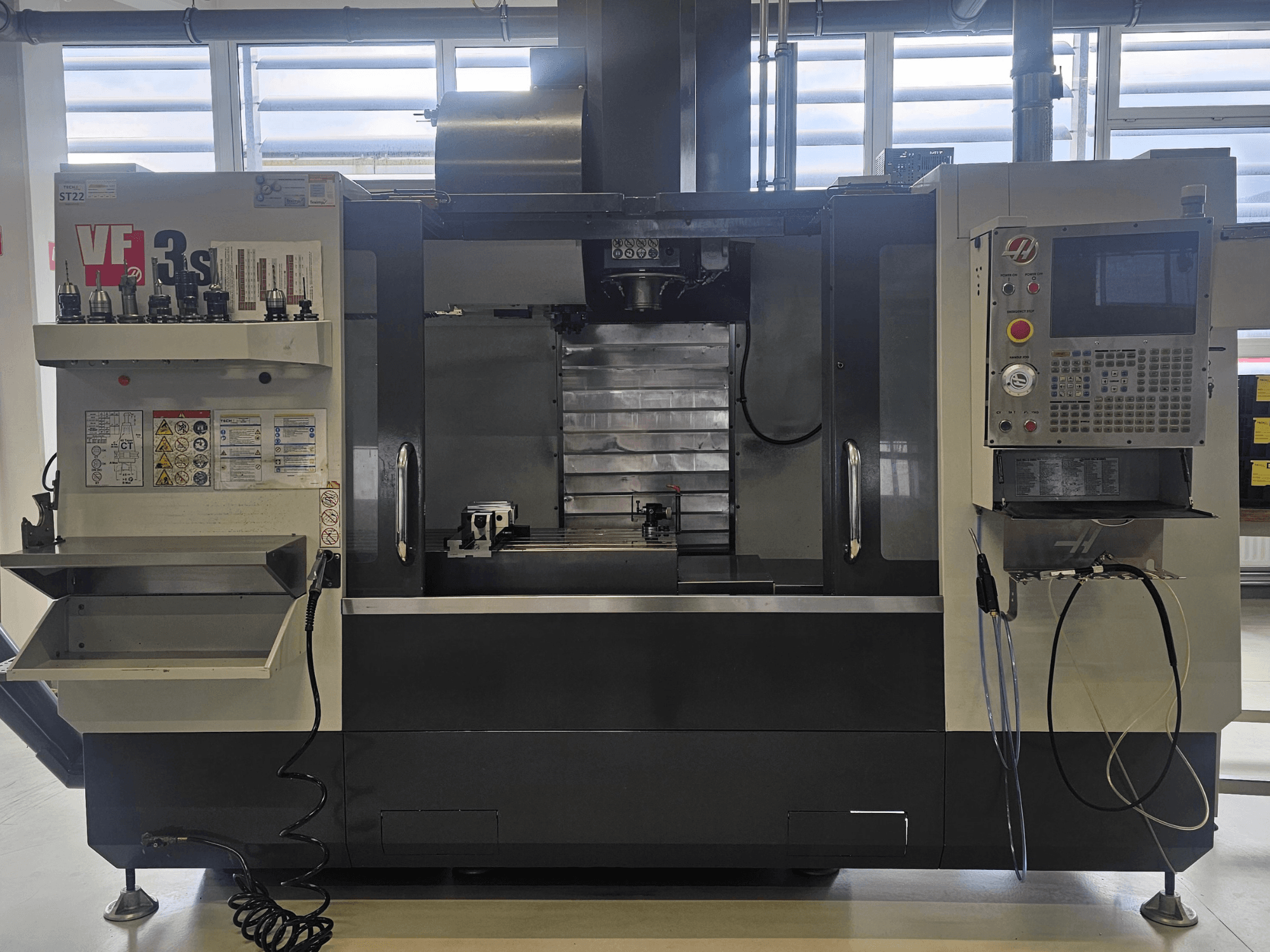 Haas VF3SS CNC frezavimo staklės, vaizdas iš priekio, įrankių laikikliai, valdymo skydelis ir tuščia aušinimo skysčio dėtuvė, matomos saugos etiketės.