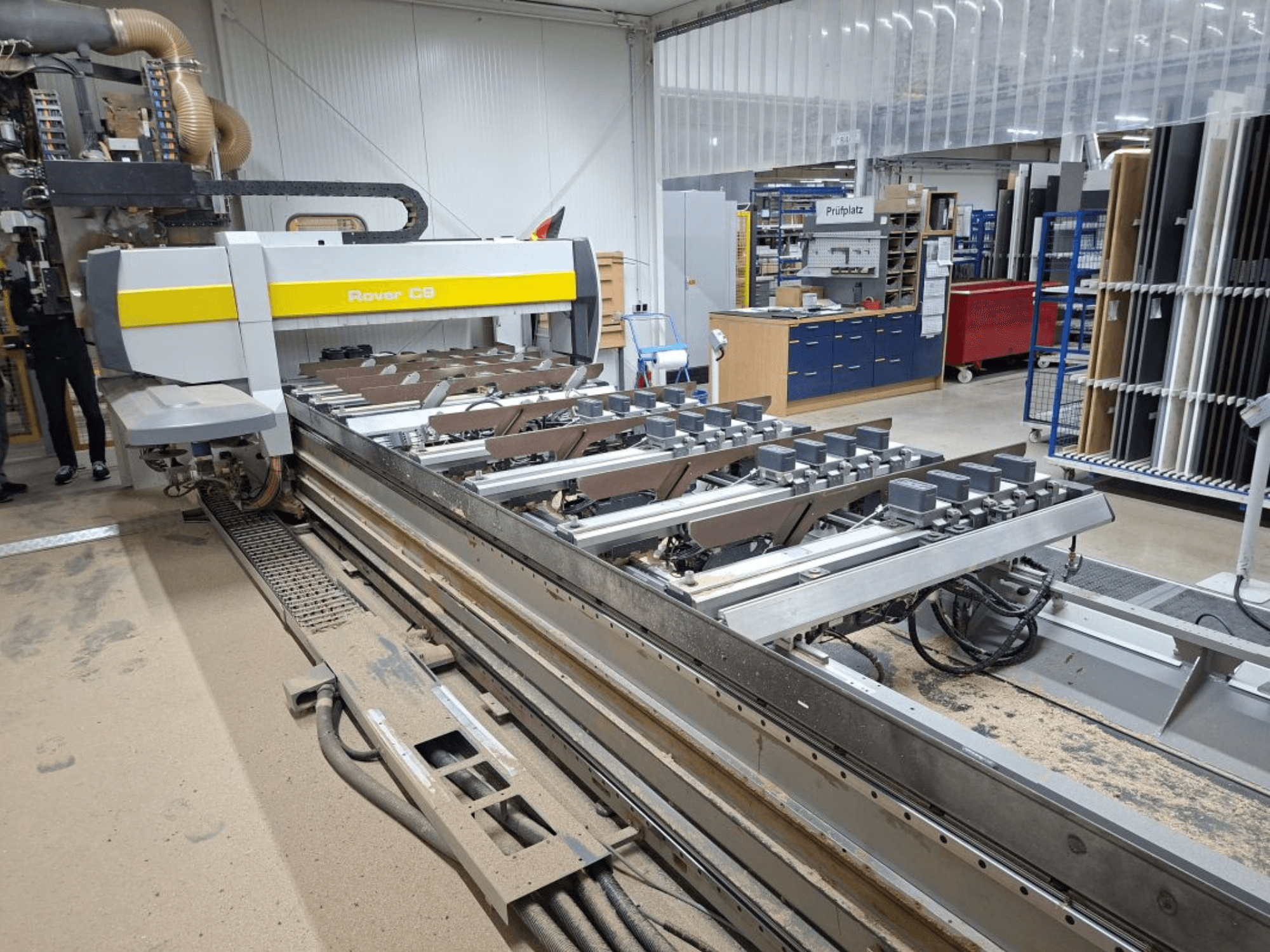 "Rover C9 CNC" staklės dirbtuvėse; iš priekio matyti pjovimo įrankiai ir konvejerio sistema, ant grindų kaupiasi dulkės.