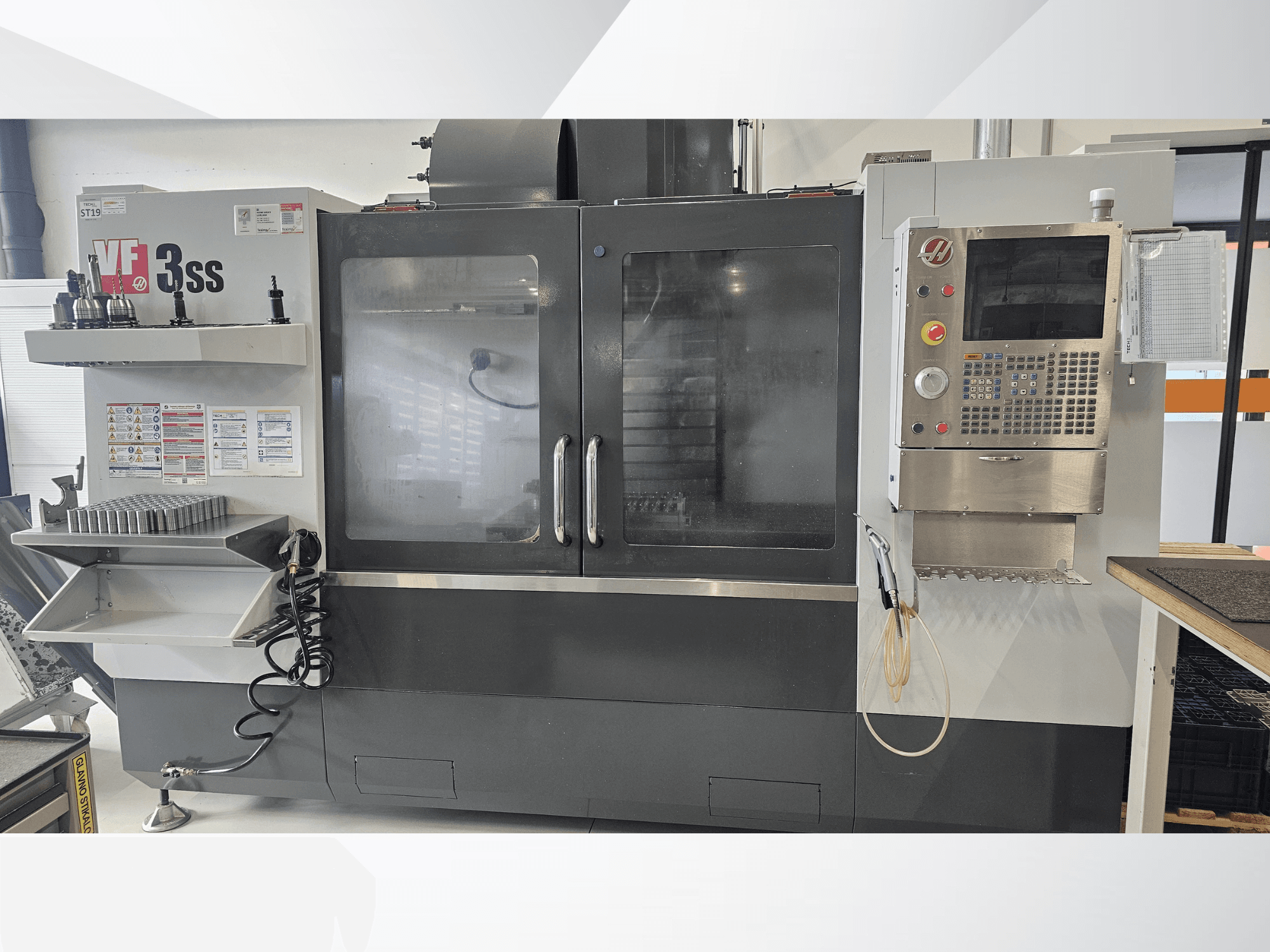 HAAS VF-3SS vertikalusis apdirbimo centras, vaizdas iš priekio, su valdymo pultu, įrankių lentyna ir didelėmis stiklinėmis durimis.