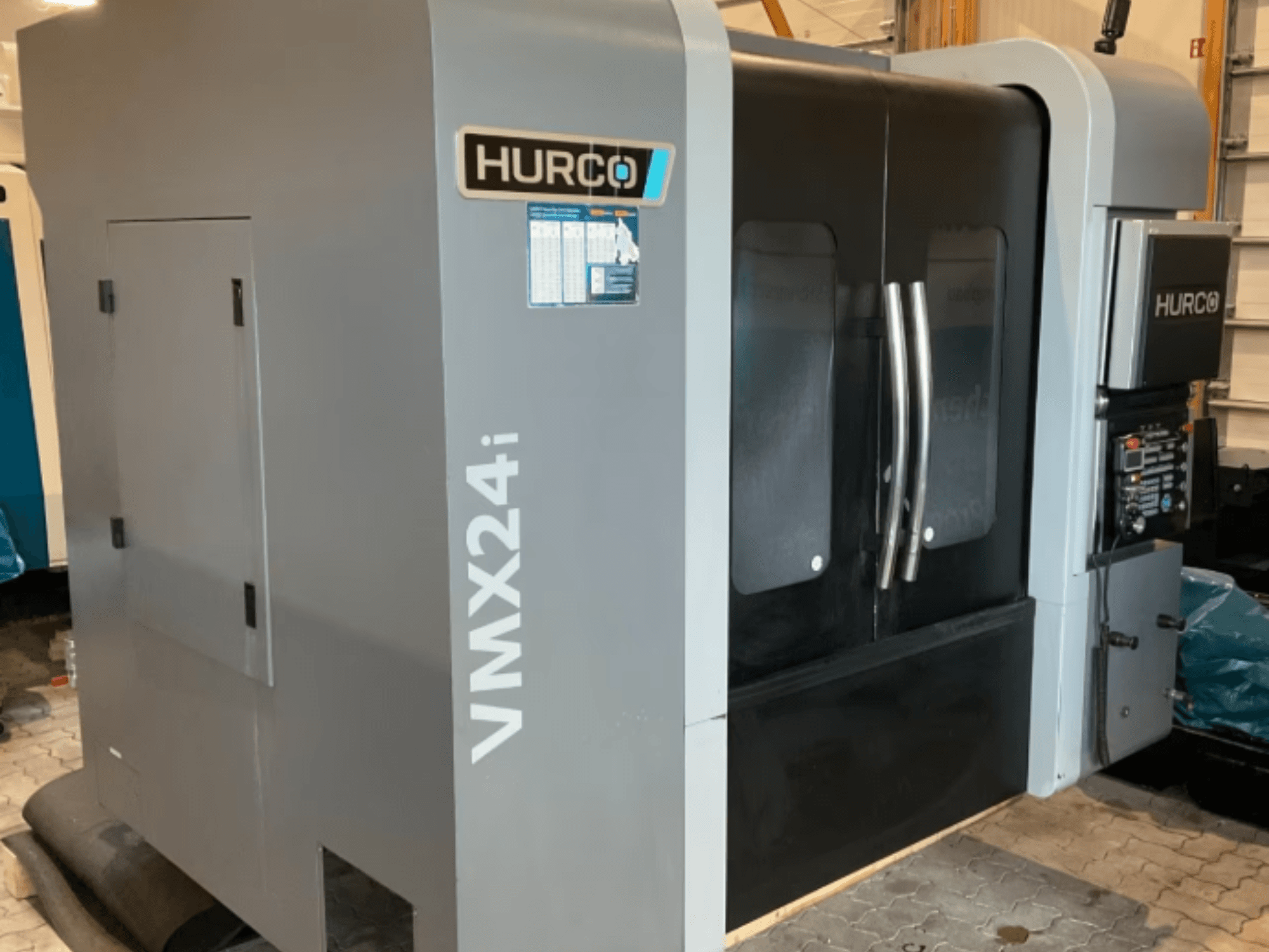 Vertikalusis apdirbimo centras „Hurco VMX 24i“, vaizdas iš priekio: metalinio pilkos spalvos korpusas, juodos spalvos skydų durelės ir valdymo pultas.