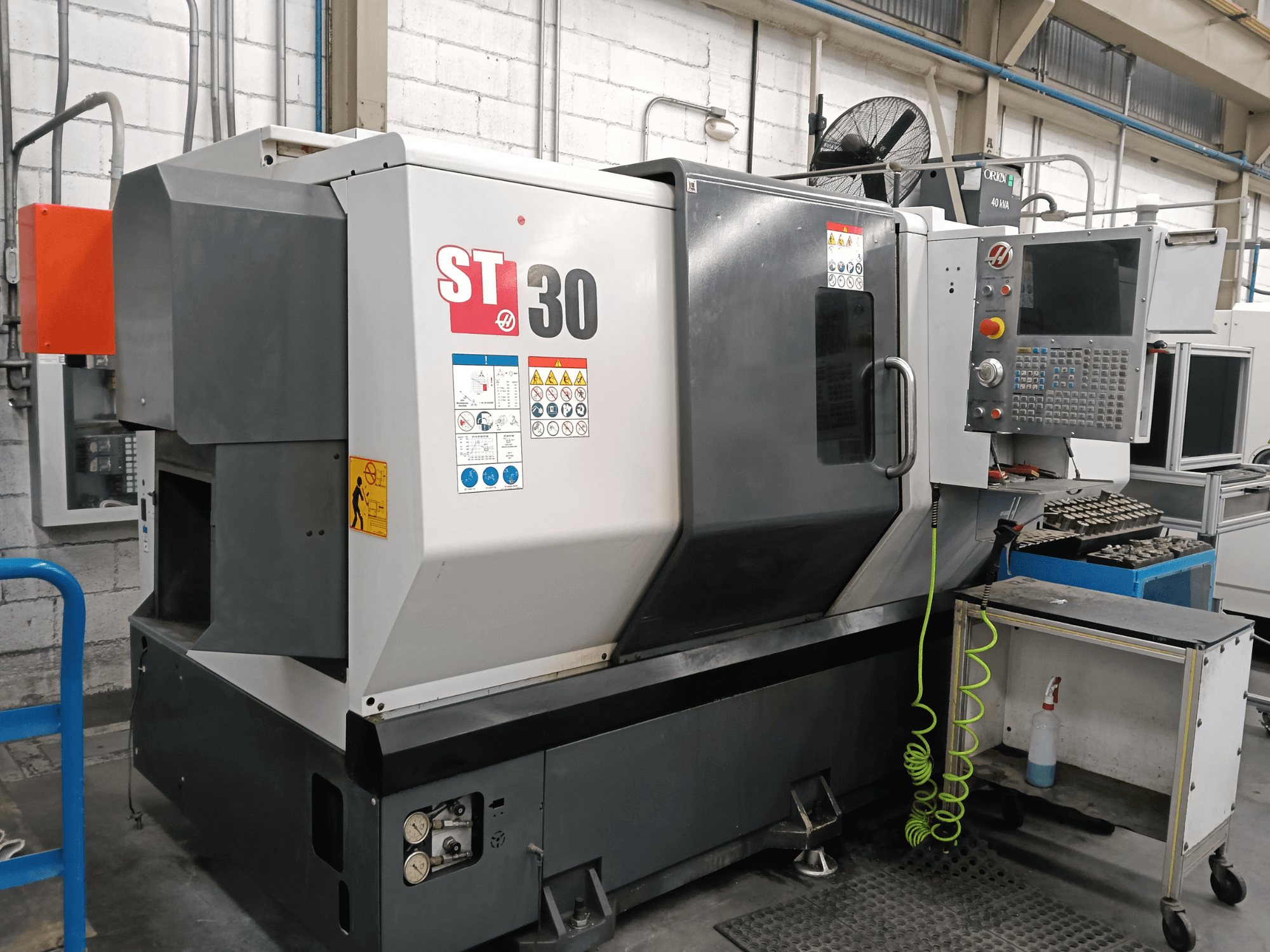 Alternatyvusis tekstas: „HAAS ST-30“ horizontalios tekinimo staklių vaizdas iš priekio su valdymo pultu, saugos etiketėmis ir įrankiais, esančiais šalia stovinčiame vežimėlyje.