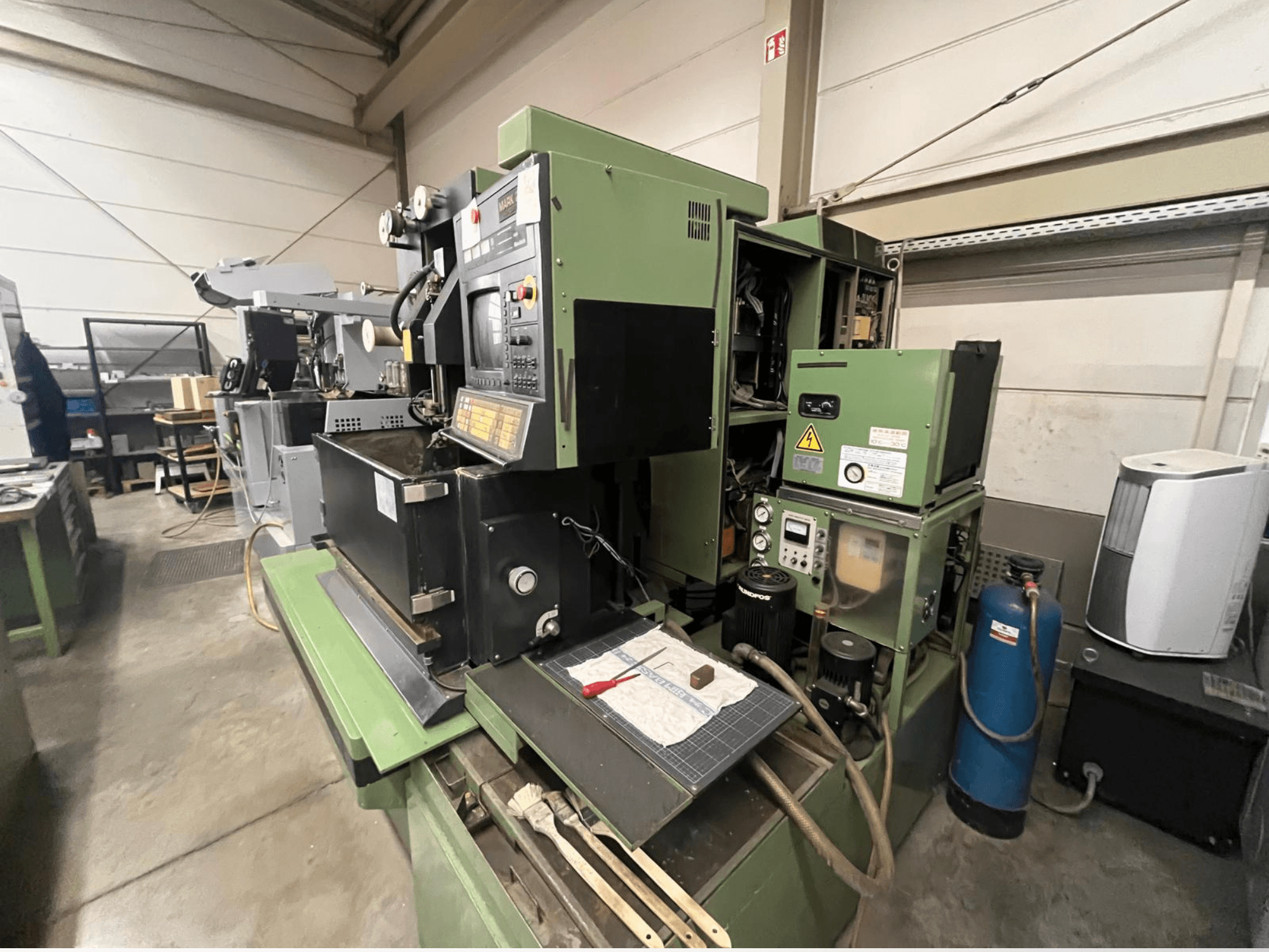 Žalios spalvos pramoninė mašina su valdymo skydeliu, vaizdas iš šono; apima hidraulinius komponentus ir darbo vietos paviršių su įrankiais.