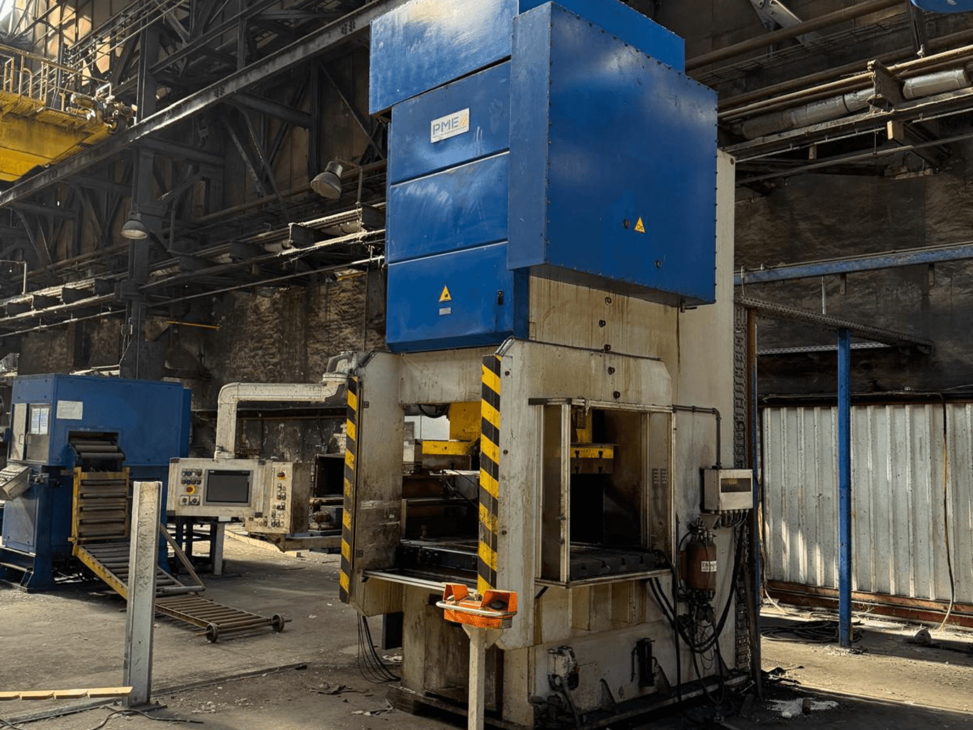 Mechaninis štampavimo presas „PME PE 400-1700 C“, vaizdas iš priekio, kuriame matyti mėlynas viršus ir geltonomis juostelėmis papuoštas pagrindas gamyklos aplinkoje.
