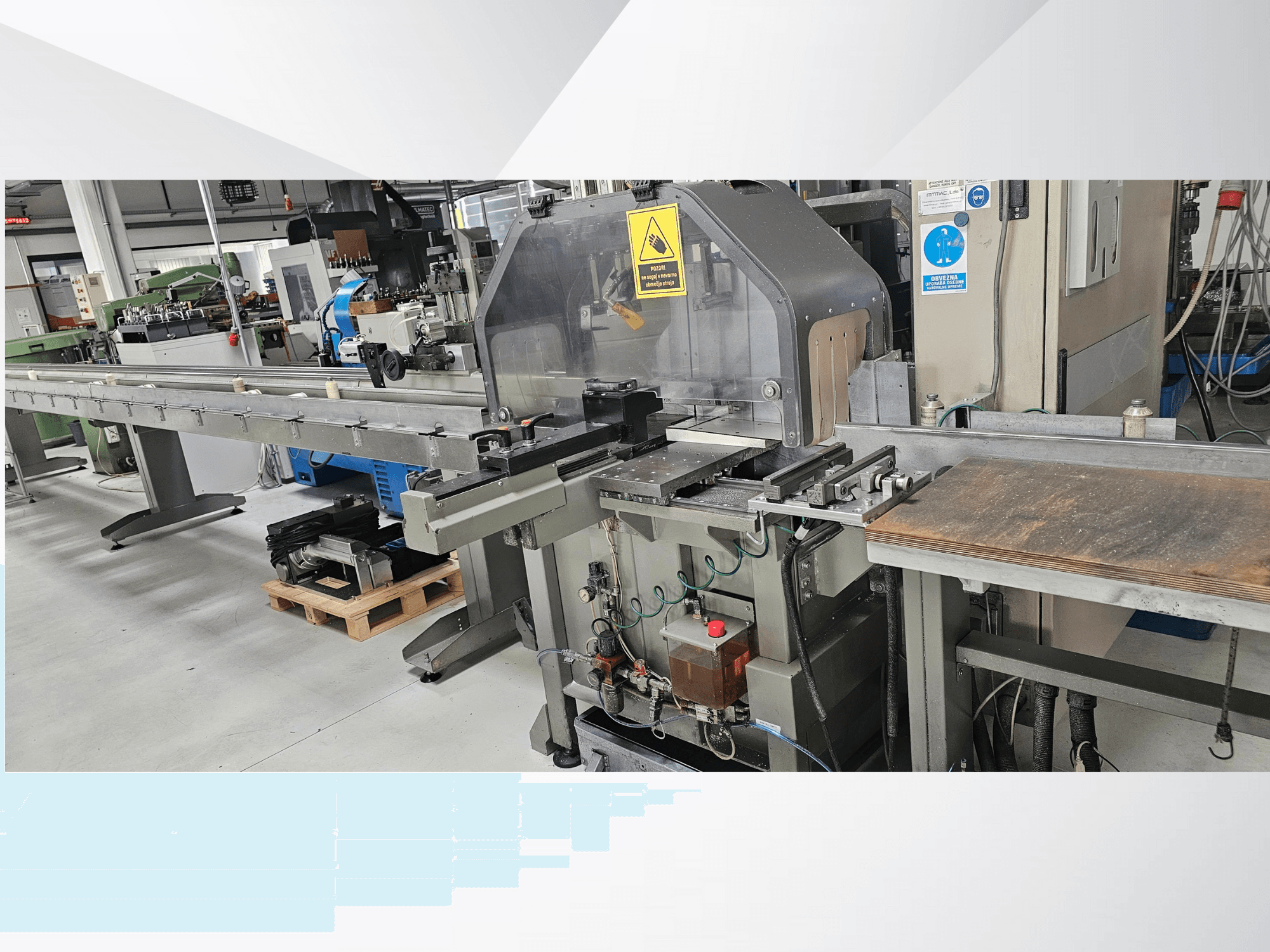 Pramoninės pjaustymo staklės iš šono, su ilgu konvejeriu, valdymo skydeliu ir saugos įspėjimais.