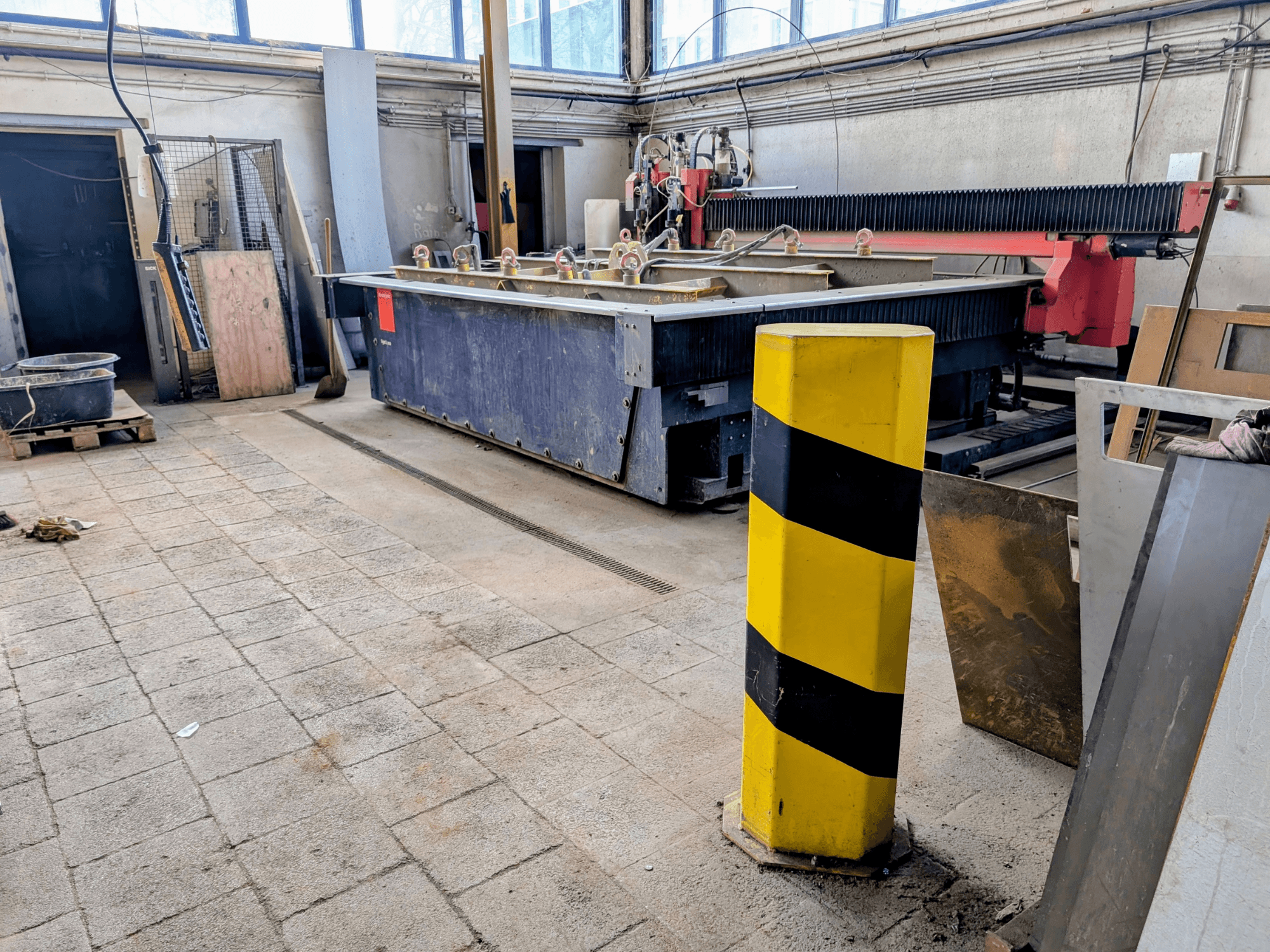 "Bystronic Byjet L 2030" pjovimo vandens srove staklės dirbtuvėse, vaizdas iš šono, su geltonu saugos stulpu ir aplinkinėmis medžiagomis.