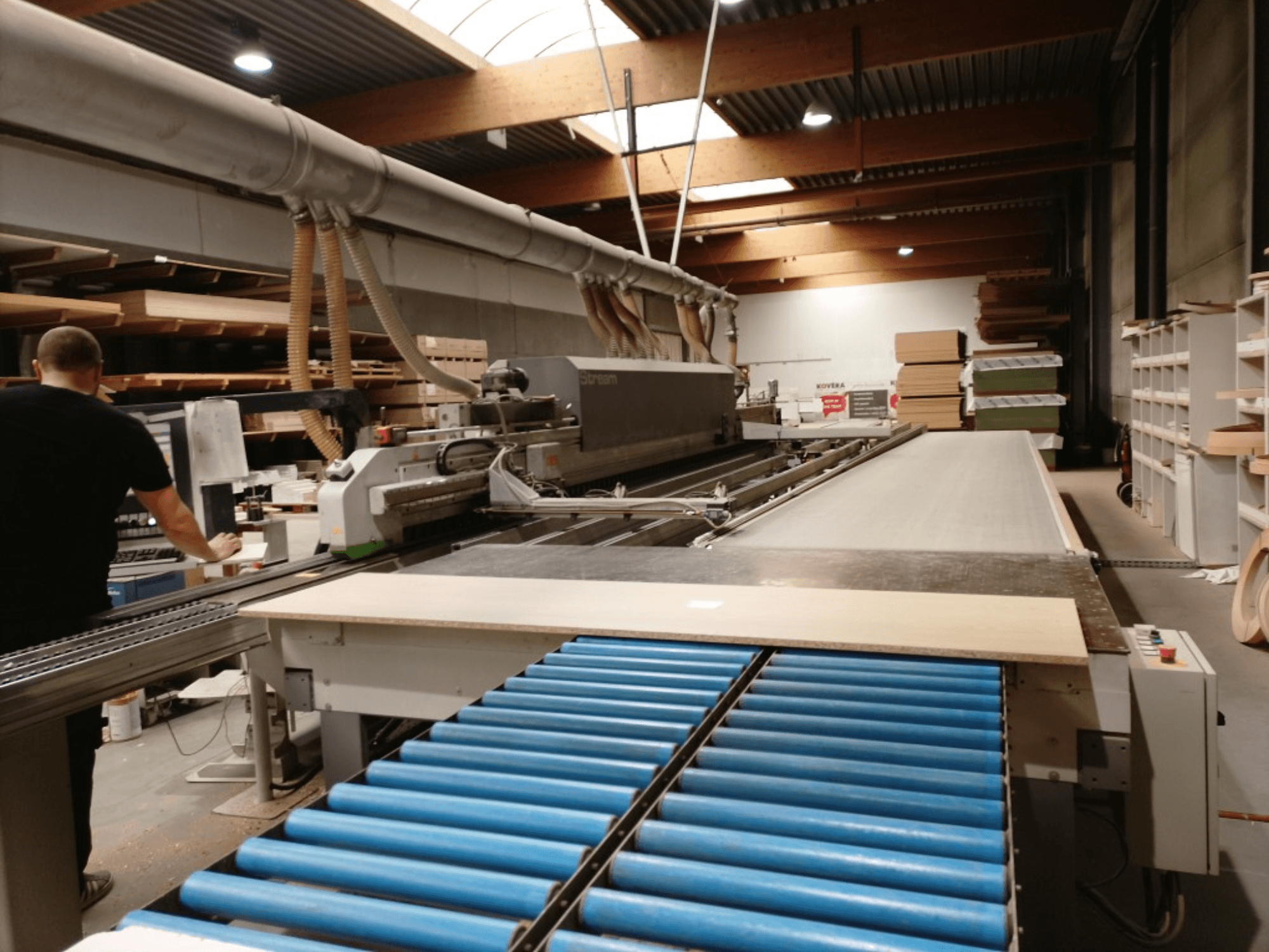 "BIESSE Edge Stream B1" kraštų briaunų apdirbimo staklės, šoninis vaizdas, kuriame matomas operatorius, mėlyni ritinėliai ir medienos plokštė ant transporterio.