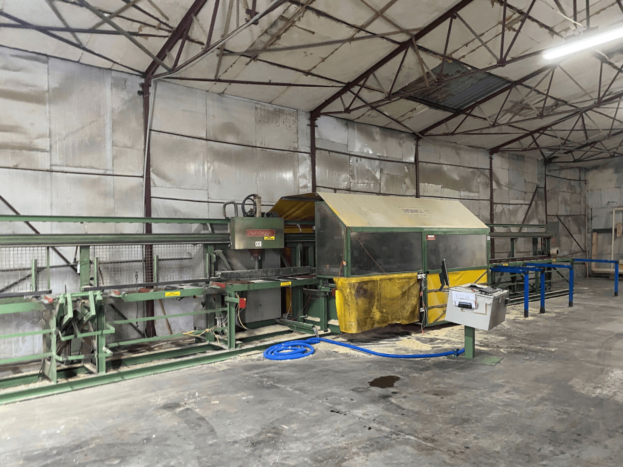 CNC medienos apdirbimo centras HUNDEGGER K1 dirbtuvėse, šoninis vaizdas, kuriame matyti mašinos, valdikliai ir pjuvenos ant grindų.