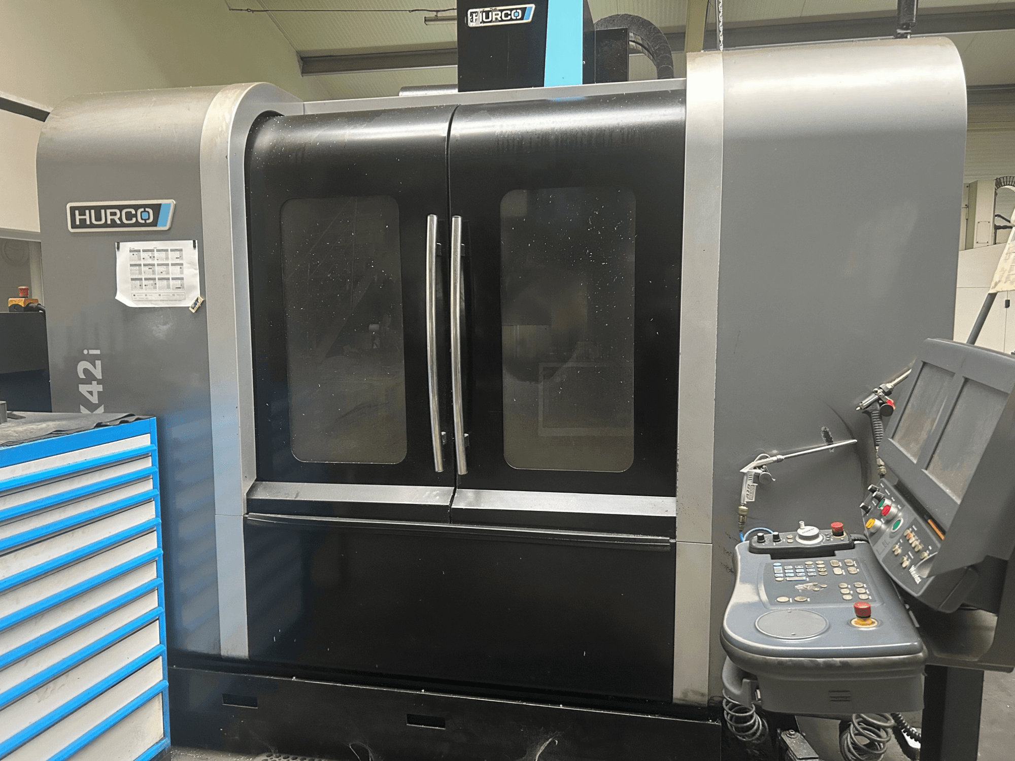 Hurco VMX42i CNC staklės, vaizdas iš priekio su valdymo skydeliu šone, dvigubos durys ir matomos techninės detalės.