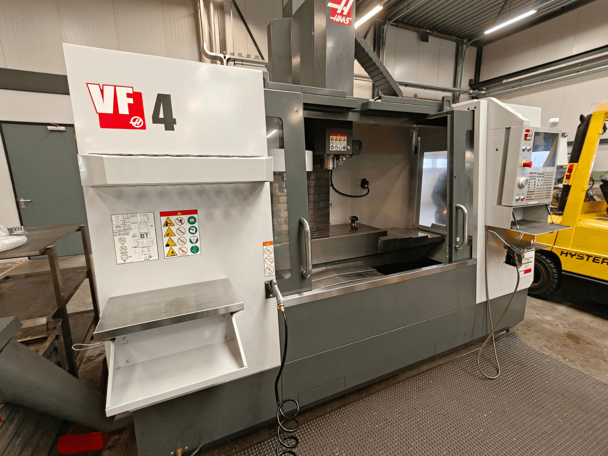 HAAS VF-4 vertikalusis apdirbimo centras, vaizdas iš priekio, su valdymo skydeliu, darbo zona ir saugos etiketėmis šone.