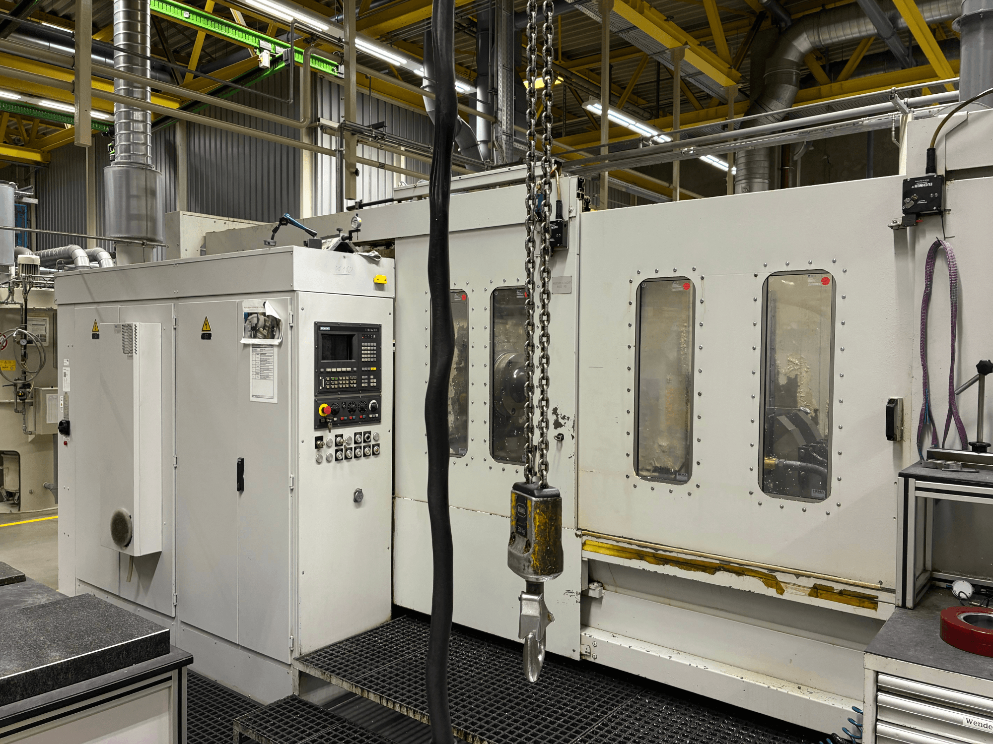 Pramoninės CNC staklės su valdymo skydeliu, vaizdas iš šono; langai, grandinės ir metaliniai komponentai gamykloje.