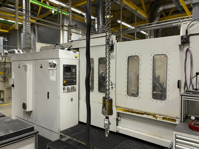 Pramoninės CNC staklės su valdymo skydeliu, vaizdas iš šono; langai, grandinės ir metaliniai komponentai gamykloje.