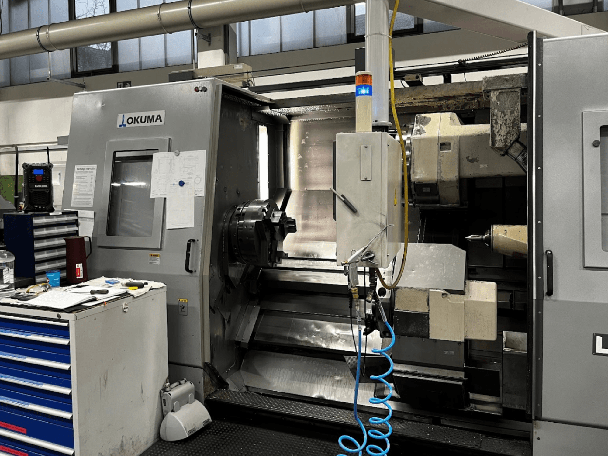 Okuma LU35-M horizontalios tekinimo staklės, vaizdas iš priekio. Valdymo pultas, įrankiai ir didelis griebtuvas, skirtas apdirbimo operacijoms.